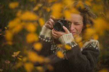 Beyaz kadın fotoğrafçı, sarı bir sonbahar ormanında eski model bir film kamerasıyla fotoğraf çekiyor. Kameralı yetişkin bir kadın.