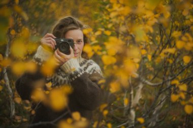 Beyaz kadın fotoğrafçı, sarı bir sonbahar ormanında eski model bir film kamerasıyla fotoğraf çekiyor. Kameralı yetişkin bir kadın.