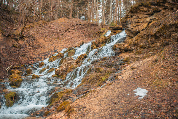 A stone dolomite waterfall with a strong stream of water flowing down. There is a forest nearby