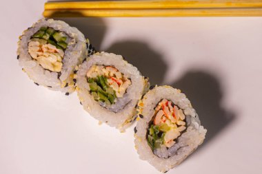 Nori yaprakları, pilav, salatalık, somon balığı suşi çeşitleri. Sushi, yemek çubuklarıyla beyaz bir arka plan tabağında yuvarlanıyor. Karışık seçmeli odak.