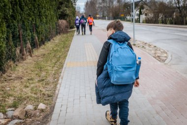 Okuldan bir çocuk özel evlerin önünden asfalt bir yol boyunca yürüyor. Bahar.