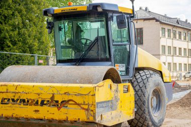 İnşaat alanına metal çitlerle çevrili sarı bir Dynapac yol silindiri yerleştirildi. Arka planda çiçek açan leylak çalıları ve yeşil yapraklar görülebilir.