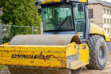 İnşaat alanına metal çitlerle çevrili sarı bir Dynapac yol silindiri yerleştirildi. Arka planda çiçek açan leylak çalıları ve yeşil yapraklar görülebilir.