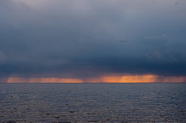 Sakin bir okyanusun üzerinde koyu, ağır bulutların altında parlak turuncu renkleri olan dramatik bir gün batımı. Ufuk, sakin su ve tehdit eden gökyüzü arasında çarpıcı bir kontrastı gösteriyor..