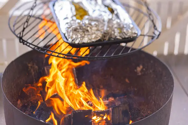 stock image Foil-wrapped food grilling over an open flame on a circular grill. The foil packets are placed in a baking tray, and flames are visible below. Perfect for outdoor cooking and barbecue enthusiasts. 