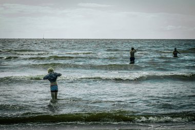 Bulutlu bir günde üç kişi okyanus dalgalarına dalıyor. Çizgili mayo ve ceketli bir çocuk kıyıya en yakın yerde dururken, iki yetişkin daha suya girmeye teşebbüs ediyor.. 