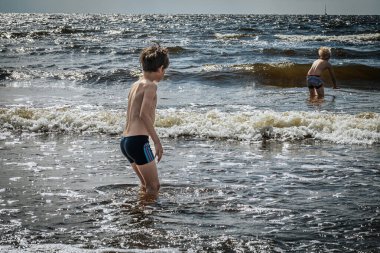 Yüzme şortlu iki çocuk, sığ okyanus dalgalarında oynayarak kumsalda güneşli bir günün tadını çıkarıyor. Bir çocuk eğilir ve diğeri suyun içinde durur. Dalgalar ve köpüklü su canlı bir zemin oluşturur.