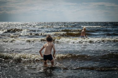 Yüzme şortlu iki çocuk, sığ okyanus dalgalarında oynayarak kumsalda güneşli bir günün tadını çıkarıyor. Bir çocuk eğilir ve diğeri suyun içinde durur. Dalgalar ve köpüklü su canlı bir zemin oluşturur.