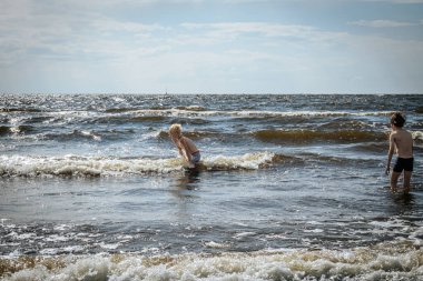 Yüzme şortlu iki çocuk, sığ okyanus dalgalarında oynayarak kumsalda güneşli bir günün tadını çıkarıyor. Bir çocuk eğilir ve diğeri suyun içinde durur. Dalgalar ve köpüklü su canlı bir zemin oluşturur.