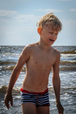 Sarı çizgili mayo giymiş genç bir çocuk kumsalda güneşli bir günün tadını çıkarıyor. Yüzünde neşeli bir ifadeyle hafifçe öne eğiliyor..
