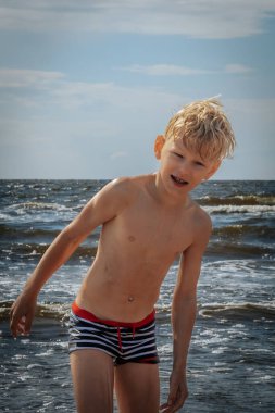 Sarı çizgili mayo giymiş genç bir çocuk kumsalda güneşli bir günün tadını çıkarıyor. Yüzünde neşeli bir ifadeyle hafifçe öne eğiliyor..