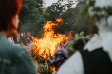 Geleneksel bir kutlama için kalabalık etrafında toplanırken büyük bir şenlik ateşi ışıl ışıl yanıyor. Sahne, ağaçların arka planına dayanır ve sıcak bir ortam yaratır.. 