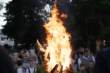 Geleneksel bir kutlama için kalabalık etrafında toplanırken büyük bir şenlik ateşi ışıl ışıl yanıyor. Sahne, ağaçların arka planına dayanır ve sıcak bir ortam yaratır.. 
