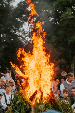 Geleneksel bir kutlama için kalabalık etrafında toplanırken büyük bir şenlik ateşi ışıl ışıl yanıyor. Sahne, ağaçların arka planına dayanır ve sıcak bir ortam yaratır.. 