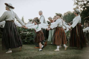 Geleneksel halk kostümleri ve çiçek taçları giymiş çocuklar ve yetişkinlerden oluşan büyük bir grup, dışarıda yeşil bir parkta toplanıyor..
