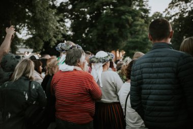 Geleneksel halk kostümleri ve çiçek taçları giymiş çocuklar ve yetişkinlerden oluşan büyük bir grup, dışarıda yeşil bir parkta toplanıyor..