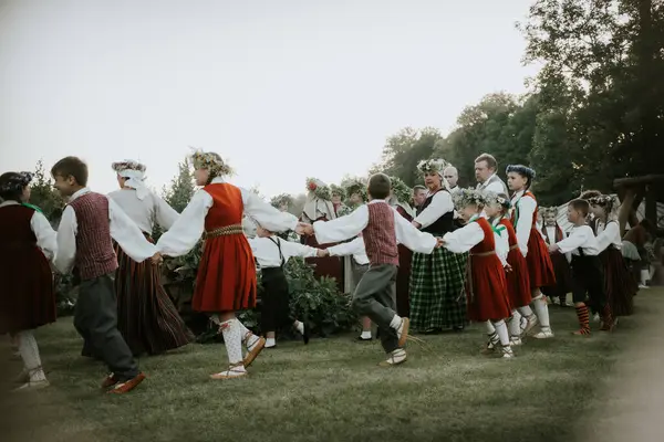 Geleneksel halk kostümleri ve çiçek taçları giymiş çocuklar ve yetişkinlerden oluşan büyük bir grup, dışarıda yeşil bir parkta toplanıyor..