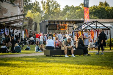 Tahta paletler ve çimenlerin üzerinde oturan bir grup insan açık havada eğleniyor. Arkaplanda resimli parlak sarı bir karatahta görülüyor.