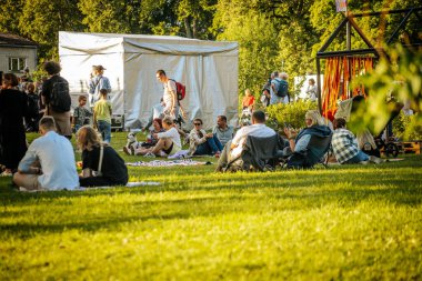 Tahta paletler ve çimenlerin üzerinde oturan bir grup insan açık havada eğleniyor. Arkaplanda resimli parlak sarı bir karatahta görülüyor.
