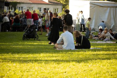 Tahta paletler ve çimenlerin üzerinde oturan bir grup insan açık havada eğleniyor. Arkaplanda resimli parlak sarı bir karatahta görülüyor.