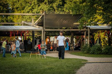 Tahta paletler ve çimenlerin üzerinde oturan bir grup insan açık havada eğleniyor. Arkaplanda resimli parlak sarı bir karatahta görülüyor.