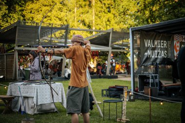 Genç bir adam bir etnik müzik etkinliğinde ok ve yayla nişan alırken, diğerleri arka planda. Sahnede masaları ve yeşillikleri olan kırsal bir ortam var.. 
