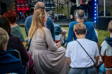 Meşgul bir seyirci bir etnik müzik festivalinde çimlerin üzerinde oturur ve etkinliği dikkatle izler. Kalabalık her yaştan insanı eğlendiriyor.