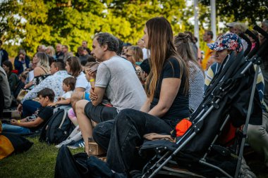 Meşgul bir seyirci bir etnik müzik festivalinde çimlerin üzerinde oturur ve etkinliği dikkatle izler. Kalabalık her yaştan insanı eğlendiriyor.