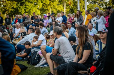 Meşgul bir seyirci bir etnik müzik festivalinde çimlerin üzerinde oturur ve etkinliği dikkatle izler. Kalabalık her yaştan insanı eğlendiriyor.