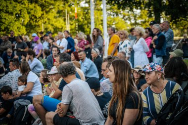 Meşgul bir seyirci bir etnik müzik festivalinde çimlerin üzerinde oturur ve etkinliği dikkatle izler. Kalabalık her yaştan insanı eğlendiriyor.