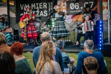 Meşgul bir seyirci bir etnik müzik festivalinde çimlerin üzerinde oturur ve etkinliği dikkatle izler. Kalabalık her yaştan insanı eğlendiriyor.