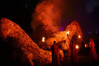  Alevli meşaleli insan figürleri aydınlatılmış bir jeodezik kubbenin yanında duruyor ve geceleri tahta bir küreden duman yükseliyor..