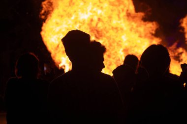 Siluetli figürler alevle yüz yüze duruyorlar, yüzleri yoğun alevlerle aydınlanıyor. Dramatik sahne, karanlık ve aydınlık arasındaki zıtlığı yakalar..