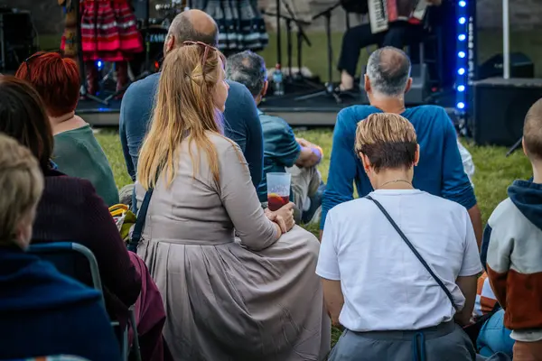 Meşgul bir seyirci bir etnik müzik festivalinde çimlerin üzerinde oturur ve etkinliği dikkatle izler. Kalabalık her yaştan insanı eğlendiriyor.