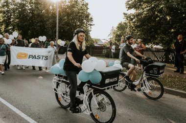 Bir grup insan kentteki farklı işletmelerin geçit töreninin yanında yürüyorlar. Sahne, dekore edilmiş araçlar ve katılımcılarla birlikte kentteki etkinliğe katılan halkın birlik ve kutlamalarını temsil ediyor..
