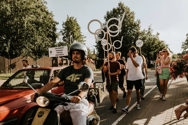 Bir grup insan kentteki farklı işletmelerin geçit töreninin yanında yürüyorlar. Sahne, dekore edilmiş araçlar ve katılımcılarla birlikte kentteki etkinliğe katılan halkın birlik ve kutlamalarını temsil ediyor..