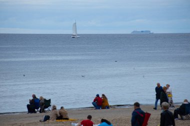 Jrmala 'da deniz kenarında görünen teknelerle yürüyen ve Letonya' nın bu kıyı şehrindeki kentsel ve doğal elementlerin karışımını yakalayan insanların yer aldığı resim gibi bir manzara..