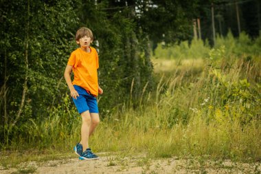Turuncu gömlekli ve mavi şortlu genç bir çocuk ağaçlık bir alanda uzun otların ve gururlu yeşilliklerin arasında duruyor. Yüzündeki ifade merakı ve oyunculuğu gösteriyor. Açık havanın ruhunu somutlaştırıyor..
