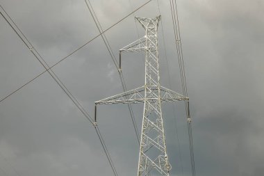 Uzun bir metal güç hattı kulesi, karanlık, bulutlu bir zemin üzerinde duruyor ve ortamı hazırlıyor. Görüntü, endüstriyel yapı ile doğal ve hareketli gökyüzü arasındaki zıtlığı vurguluyor..