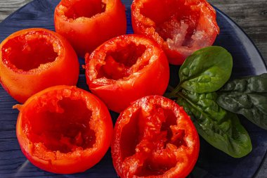 Close-up of hollowed red tomatoes ready for stuffing placed next to fresh spinach leaves on a blue plate. Tomatoes are prepared for filling. clipart