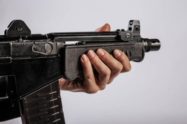 Siyah bir makineli tüfeğin ön sapını kavrayan bir elin yakın çekimi silahın vücudunun detaylarını gösteriyor. Görüntünün minimalist bir arka planı var, kopyalama alanı sağlıyor. Görünür bir logo yok.