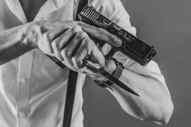 Aynı anda hem tabanca hem de bıçak tutan bir adamın siyah beyaz görüntüsü dramatik ve yoğun bir atmosfer yaratıyor. Adam pantolon askısı olan beyaz bir gömlek giyiyor, retro ya da noir ekliyor. 