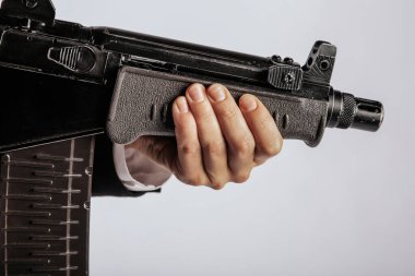 Siyah bir makineli tüfeğin ön sapını kavrayan bir elin yakın çekimi silahın vücudunun detaylarını gösteriyor. Görüntünün minimalist bir arka planı var, kopyalama alanı sağlıyor. Görünür bir logo yok.