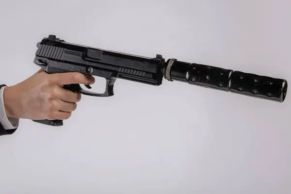 Önünde susturucu olan siyah bir tabanca tutan takım elbiseli bir el. Resmin, kopya alanı sunan minimalist beyaz bir arka planı vardır. Görünür bir logo ya da marka yok.