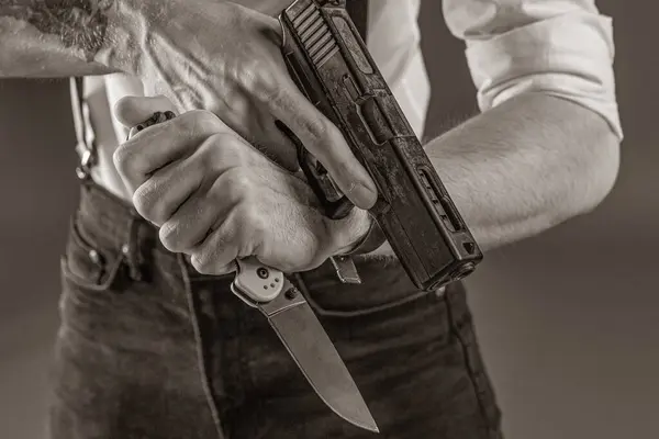 Aynı anda hem tabanca hem de bıçak tutan bir adamın siyah beyaz görüntüsü dramatik ve yoğun bir atmosfer yaratıyor. Adam pantolon askısı olan beyaz bir gömlek giyiyor, retro ya da noir ekliyor. 