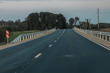 Pürüzsüz, boş bir yol bir tarafında sık ağaçlar, diğer tarafında açık alanlar olan ormanlık bir alanda esiyor. Yumuşak akşam ışığı, gökyüzünde ve üzerinde bolca fotokopi alanı olan huzurlu bir atmosfer yaratır.