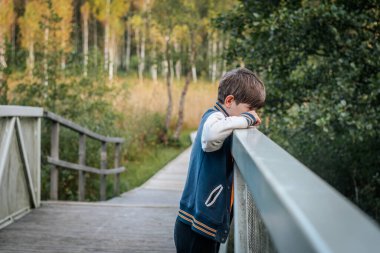 Genç bir çocuk ahşap bir köprünün korkuluğuna eğiliyor ve düşünceli bir şekilde uzaklığa bakıyor. Yeşillik ve ağaçlarla çevrili sahne, doğal ortamdaki sessiz ve düşünceli bir anı yakalar..