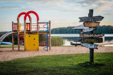 Rengarenk bir oyun alanı, yakınlardaki yerleri gösteren ahşap bir levhanın yanında kumlu bir zeminde bulunur. Arka planda sessiz bir göl ve orman, sakin bir açık hava manzarası yaratır.