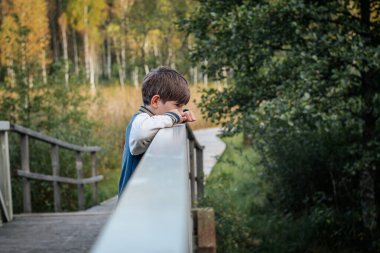 Genç bir çocuk ahşap bir köprünün korkuluğuna eğiliyor ve düşünceli bir şekilde uzaklığa bakıyor. Yeşillik ve ağaçlarla çevrili sahne, doğal ortamdaki sessiz ve düşünceli bir anı yakalar..