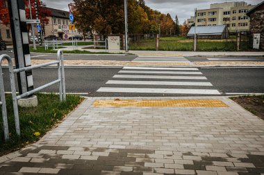 Yaya geçidi, asfalt yol boyunca geniş pencereleri olan modern bir binaya götürür. Bölge sonbahar yaprakları ve düzgün budanmış çalıları olan ağaçlarla çevrilidir. Bulutlu gökyüzü.
